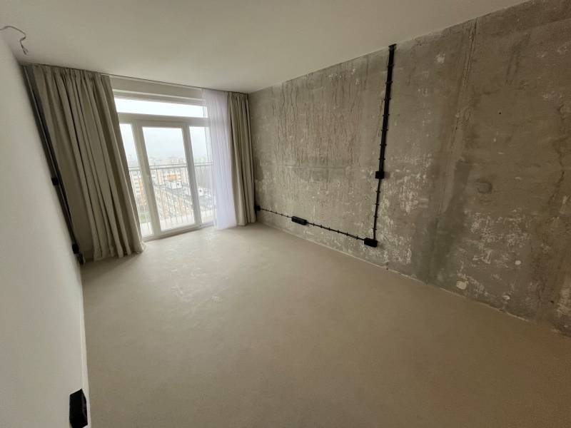 Bright studio apartment with exposed concrete, curtains, and balcony doors.