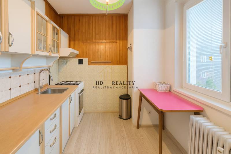A kitchen in a 3-room apartment with a wood-patterned floor, white cabinets, and a pink table.