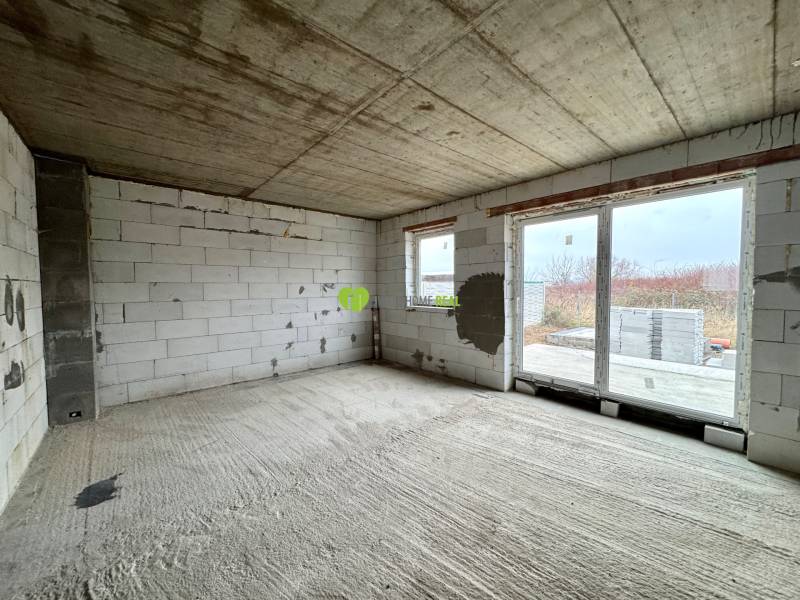 The interior of a family house with a concrete floor, raw walls, and large windows.