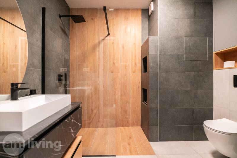A bathroom with a shower, wooden decor flooring, and dark tiles in a 3-room apartment.