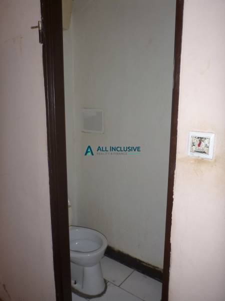 A toilet in a small two-room apartment with plain walls and ceramic tiles on the floor.
