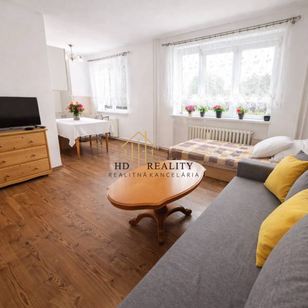 Living area in a studio apartment with a sofa, table, and wood-patterned flooring.