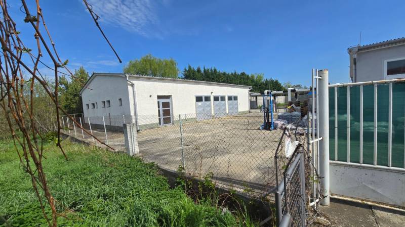 Warehouses and halls in Šaľa, a white building with a gate and fencing, surrounded by greenery.