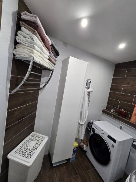 A bathroom in a 3-room apartment with a washing machine, shelves, and a wooden decor floor.