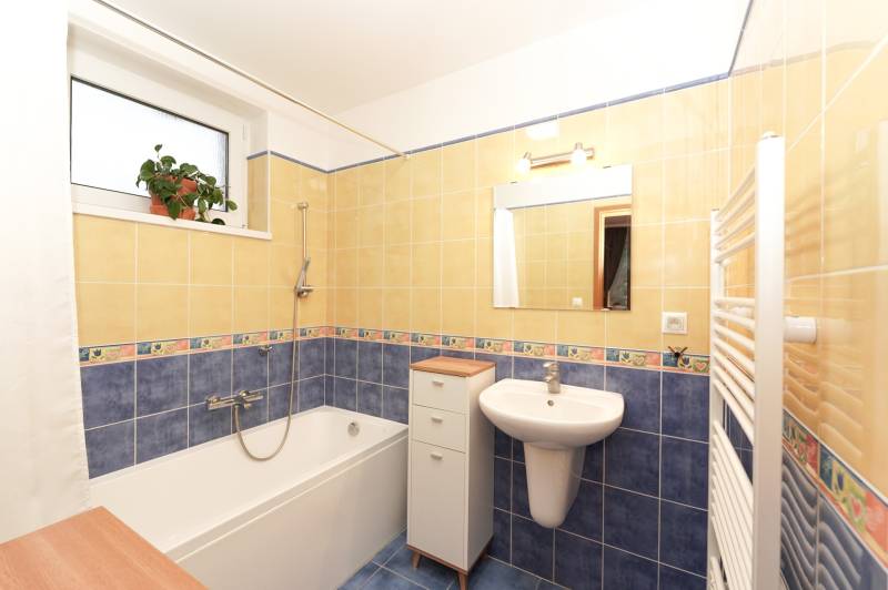 A bathroom in a family house with a bathtub, a sink, tiles, and a window with a flower.