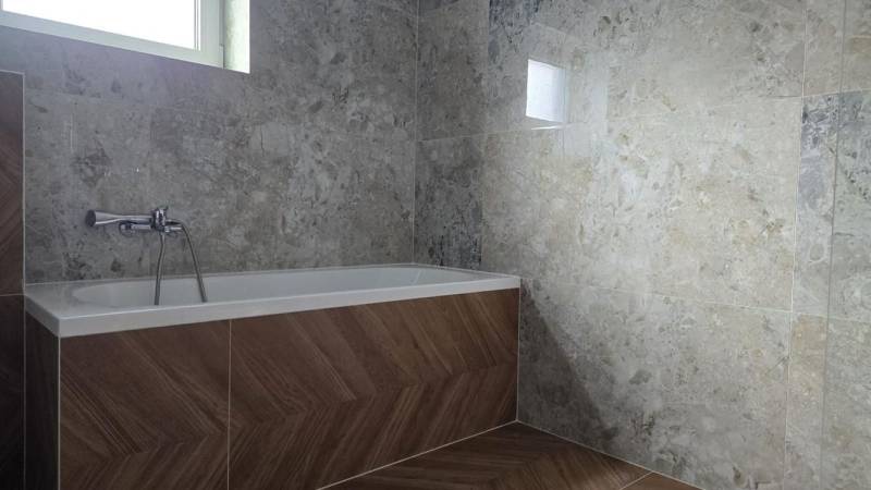 A bathroom in a family house with a bathtub, tiles with a stone pattern, and a floor with a wooden decor.