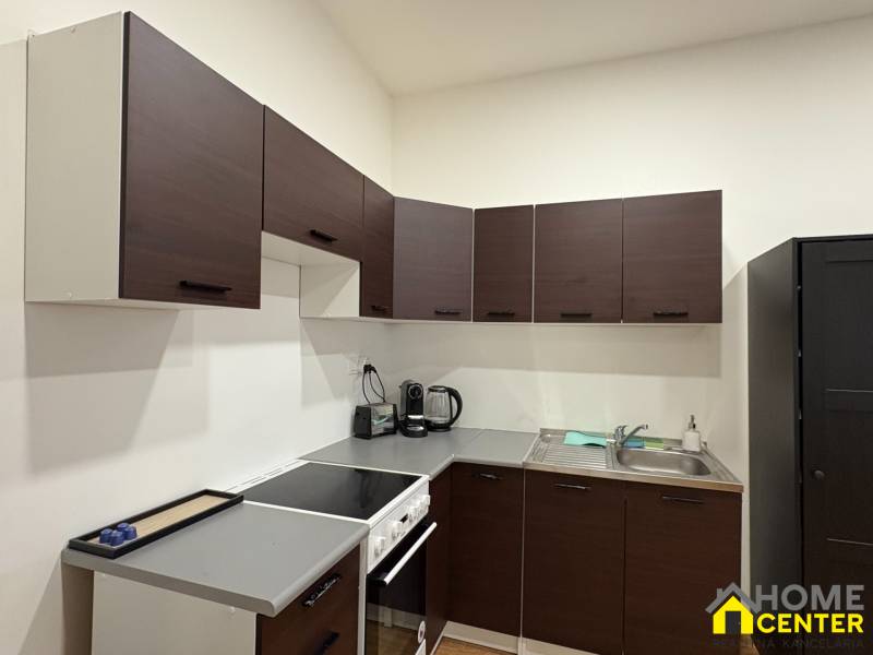 A kitchen in a studio apartment with dark cabinets and appliances, wooden decor flooring.