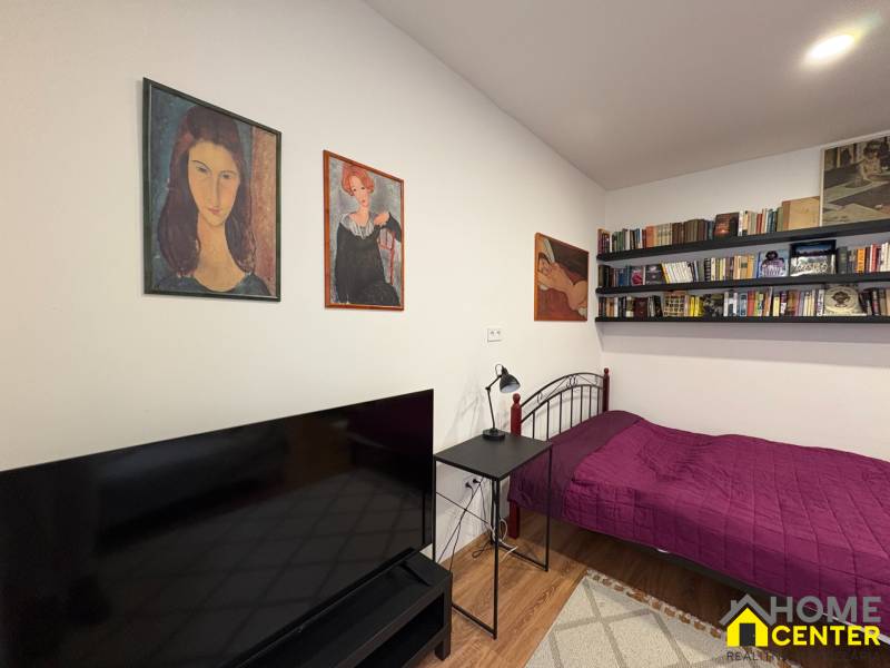 A studio apartment with a bed, bookshelves, and a television, with a wood-patterned floor.