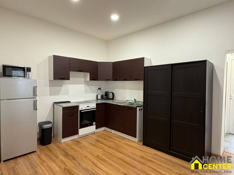 A kitchen in a studio apartment with white walls, wooden floor decor, and appliances.