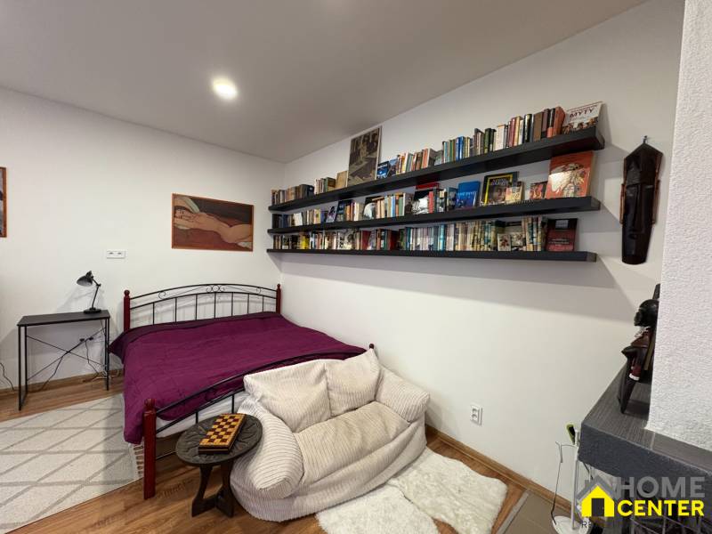 Bedroom in a one-room apartment with a bed, bookshelf, and wooden decor flooring.
