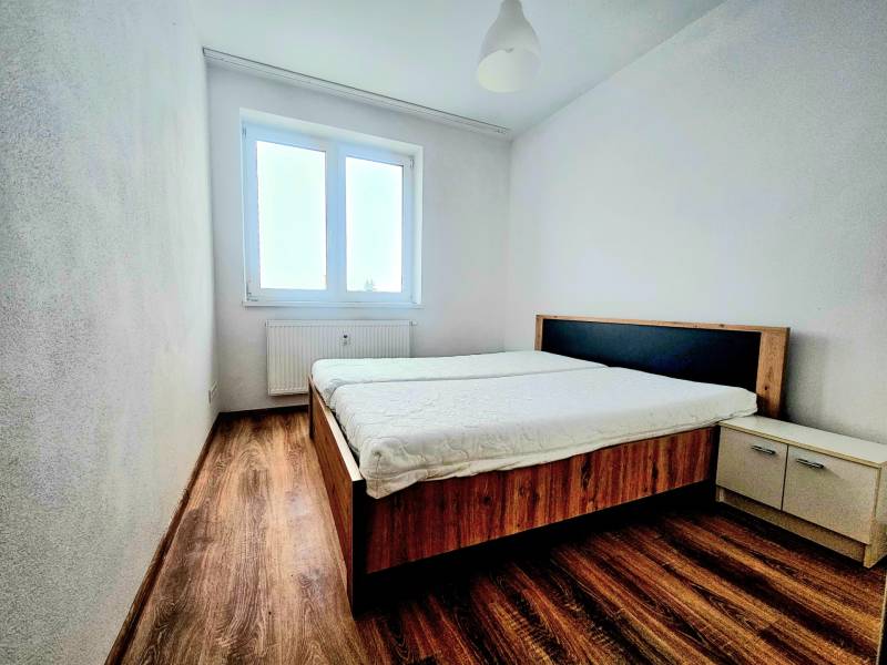 Bedroom in a two-room apartment with wood-patterned flooring, a bed, and a window.
