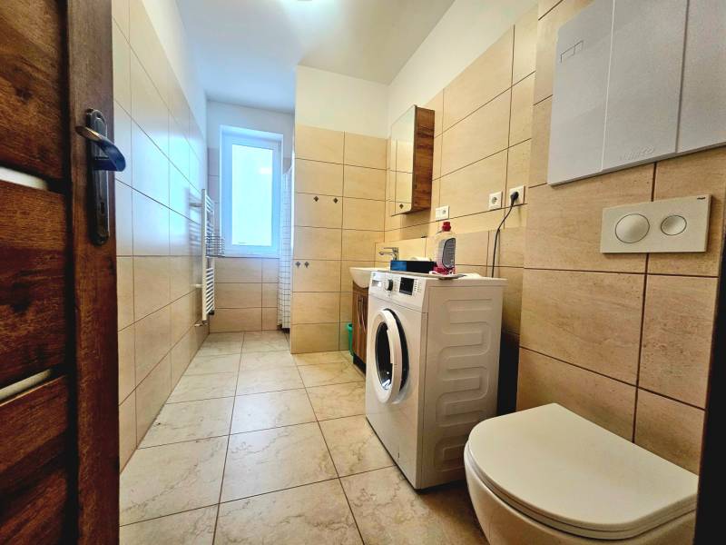 A bathroom with a washing machine and toilet in a 2-room apartment, with ceramic tiles and a bright window.