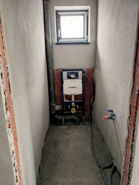 Construction preparation for a toilet in a family house with a built-in tank, white walls, and a small window.