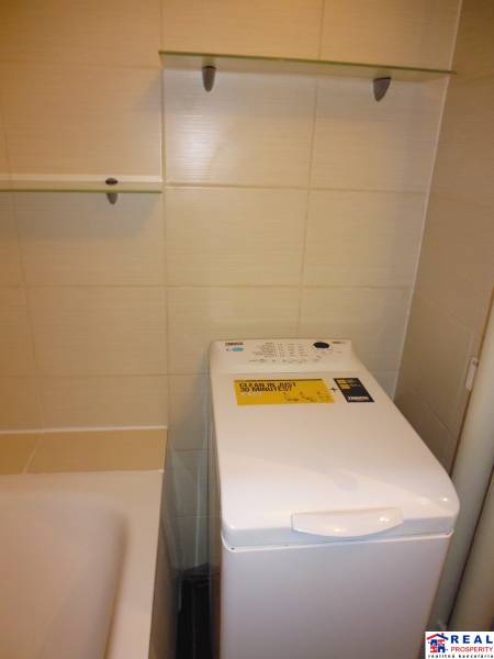 Bathroom in a 2-room apartment with a washing machine next to the bathtub and shelves.