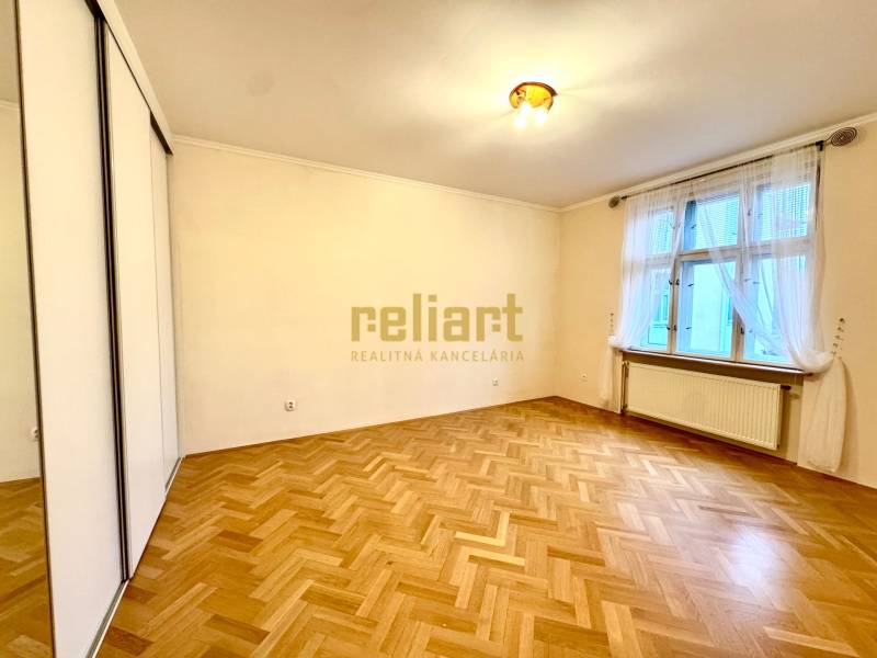 A room in a 4-room apartment with a large window, white walls, and a floor with a wood decor.