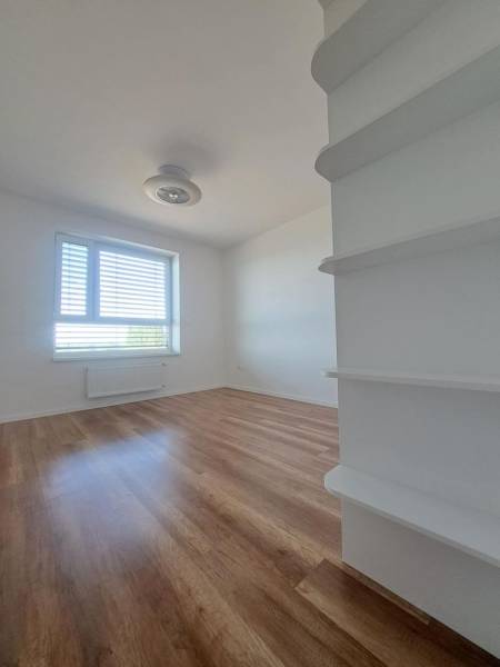 A room in a 2-room apartment with a wooden decor floor, light walls, and a window.