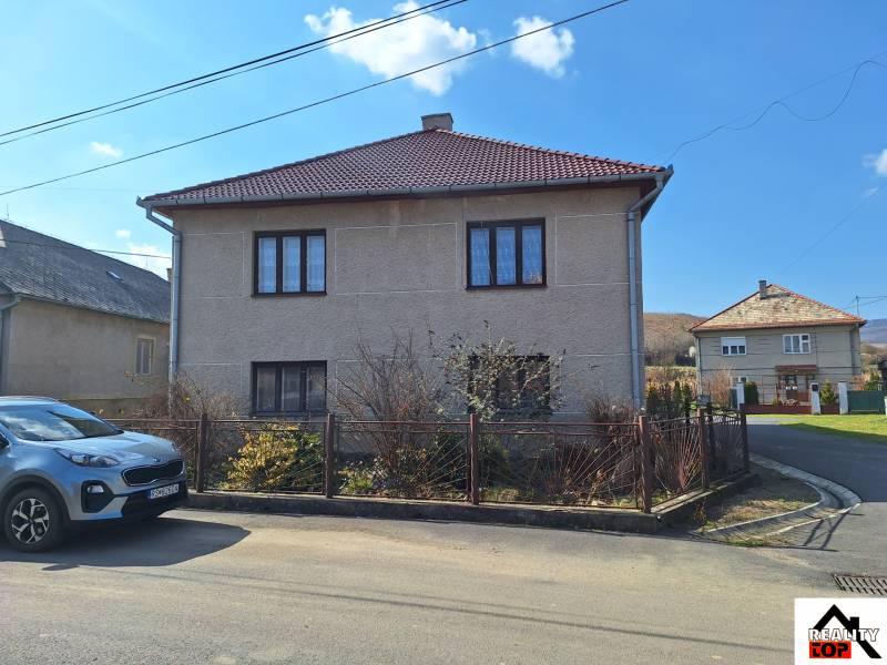 A family house in Tachtoch with a fence, a car, and a neighboring house under a blue sky.