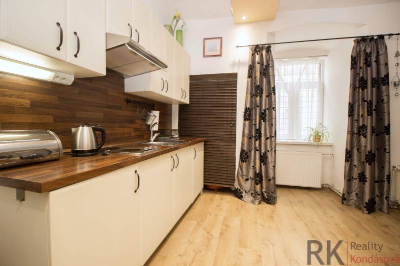 A kitchen with a wood-patterned floor in a 2-room apartment, white cabinets, and patterned curtains.