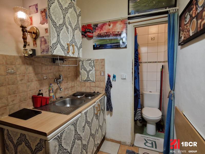 A kitchenette in a studio apartment with wallpaper and tiles, next to the toilet, the wall is decorated with photographs.