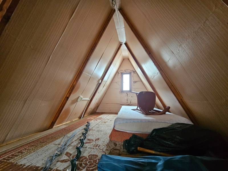 Attic of a cottage with a mattress and a chair, wooden decor, light walls.