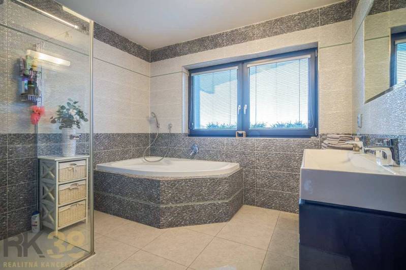 Bathroom in a family house with a bathtub, shower, and sink, light tiles, window with blinds.