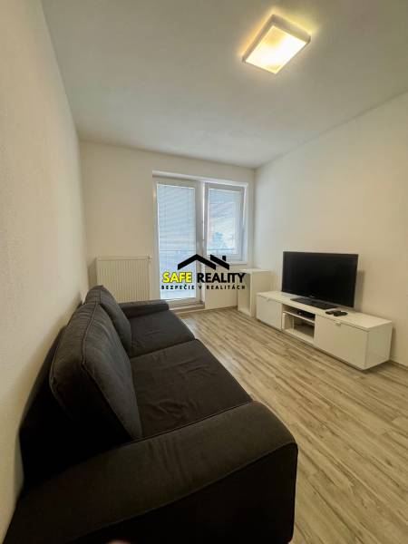 Living room in a two-room apartment with a sofa, television, and wooden decor flooring.