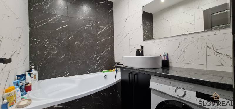 A bathroom with a bathtub, sink, and washing machine in a 2-room apartment, tiled with dark marble tiles.