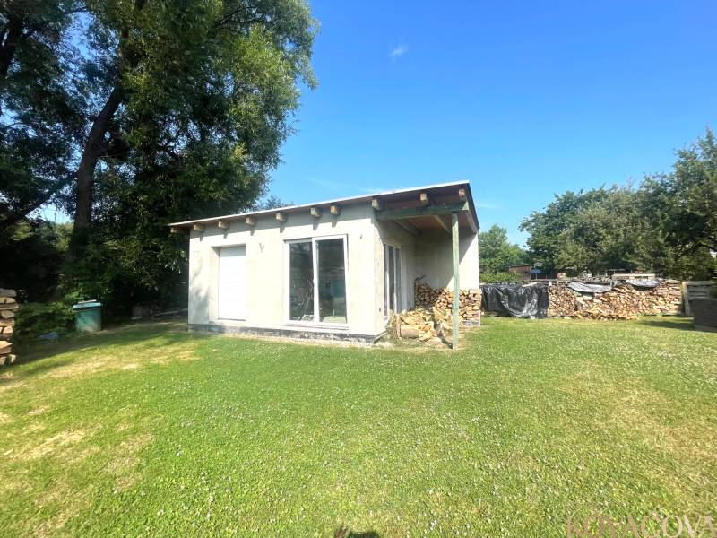 A family house in Melčice-Lieskové with a woodpile and a grassy yard surrounded by trees.