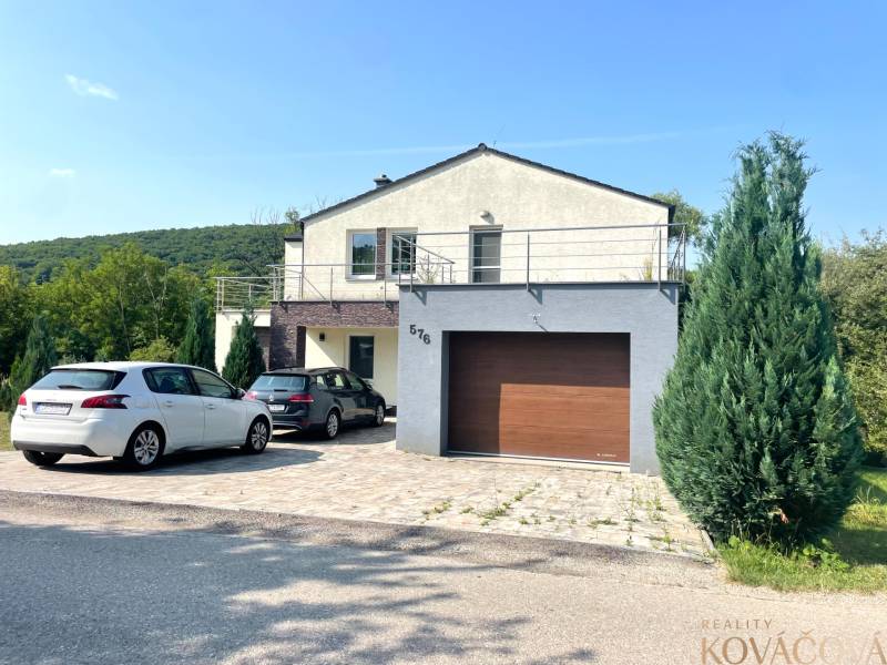 A family house in Melčice-Lieskové, with two cars parked in front, surrounded by greenery.