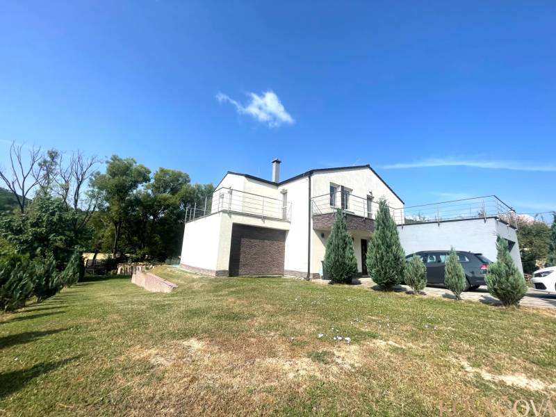 A family house in Melčice-Lieskové with a parking lot and a green lawn, surrounded by trees.