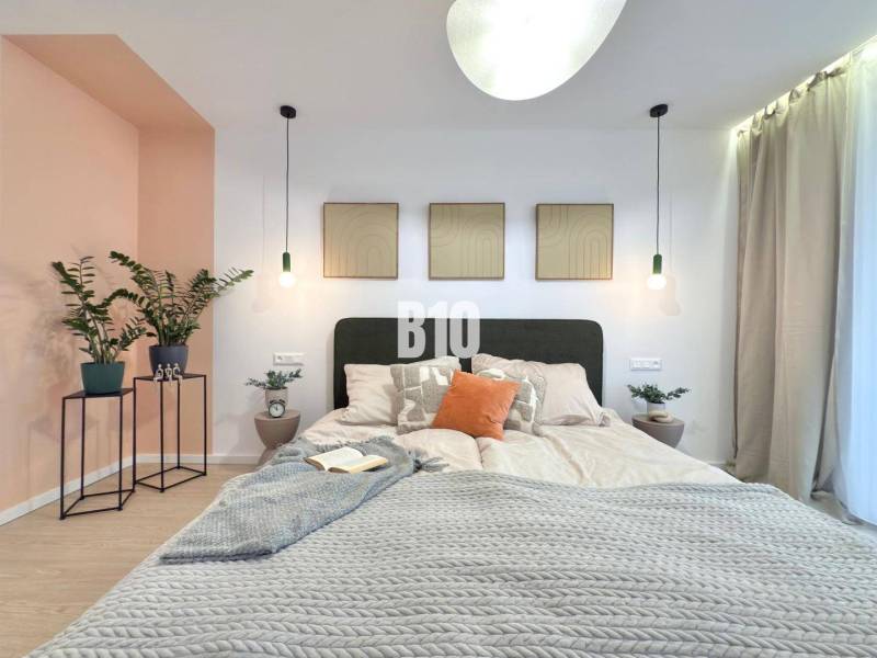 A bright bedroom with a bed, wood-patterned flooring, plants, and paintings on the wall.