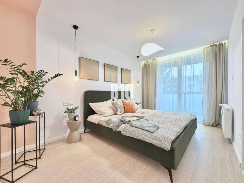 A bright bedroom with a bed and plants in a 3-room apartment with wood-patterned flooring.