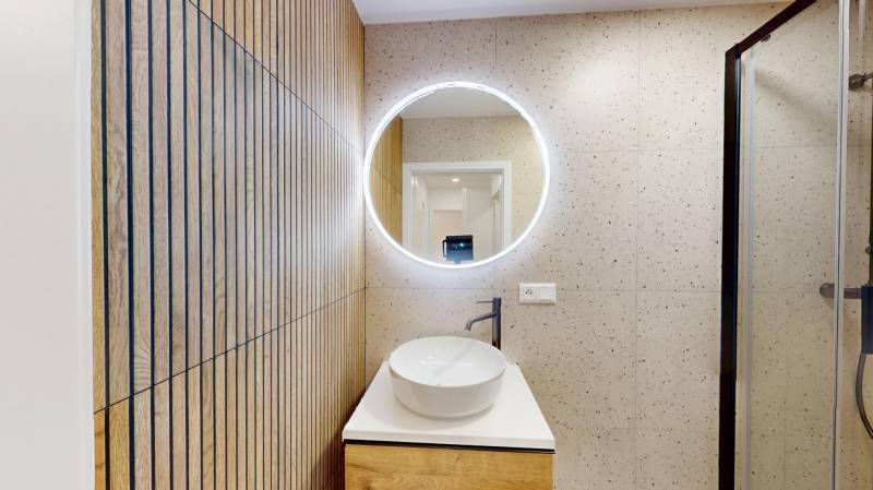 A hallway from a 4-room apartment with a sink, a mirror, and a wall with a wooden decor.