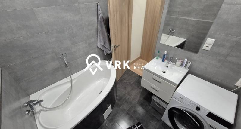Bathroom in a 2-room apartment with a bathtub, mirror, washing machine, and gray tiles.