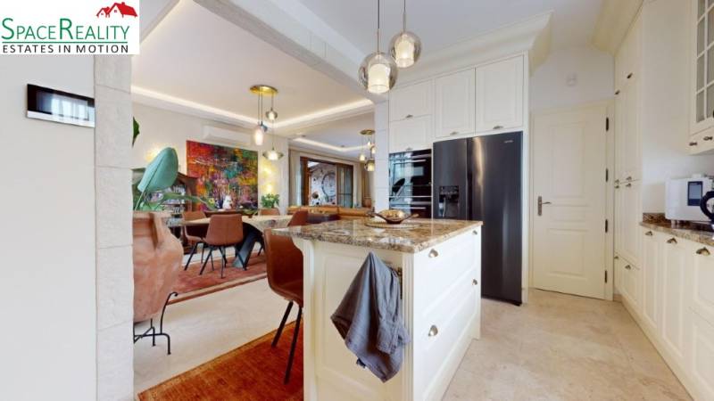 A kitchen with an island, light flooring, chairs, and artwork in a family house.