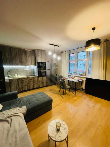 Living room with kitchen, gray sofa, wooden decor flooring, dining table, pendant lamps.