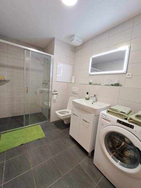 A studio apartment bathroom with an elegant shower, sink, toilet, and washing machine with green accessories.