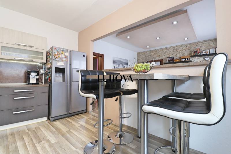 A kitchen in a 3-room apartment with bar stools, a stainless steel refrigerator, and a wooden decor floor.