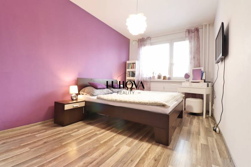 A bedroom with a light purple wall, a bed, and a wooden decor floor in a 3-room apartment.
