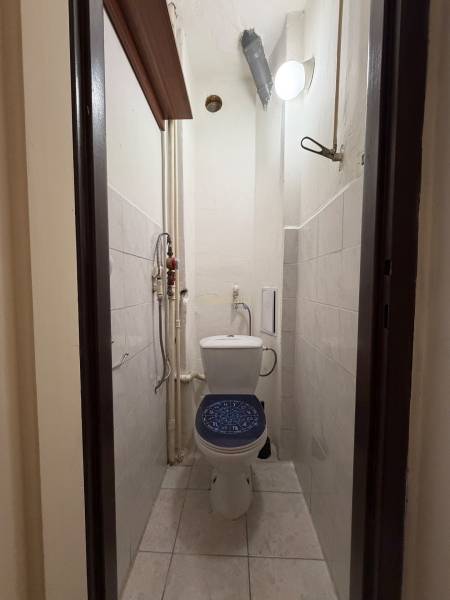 Narrow toilet with ceramic tiles and a wall light in a studio apartment.