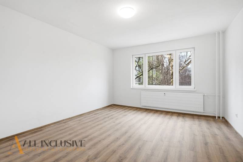 An empty living room in a three-room apartment with wood-patterned flooring and a large window.