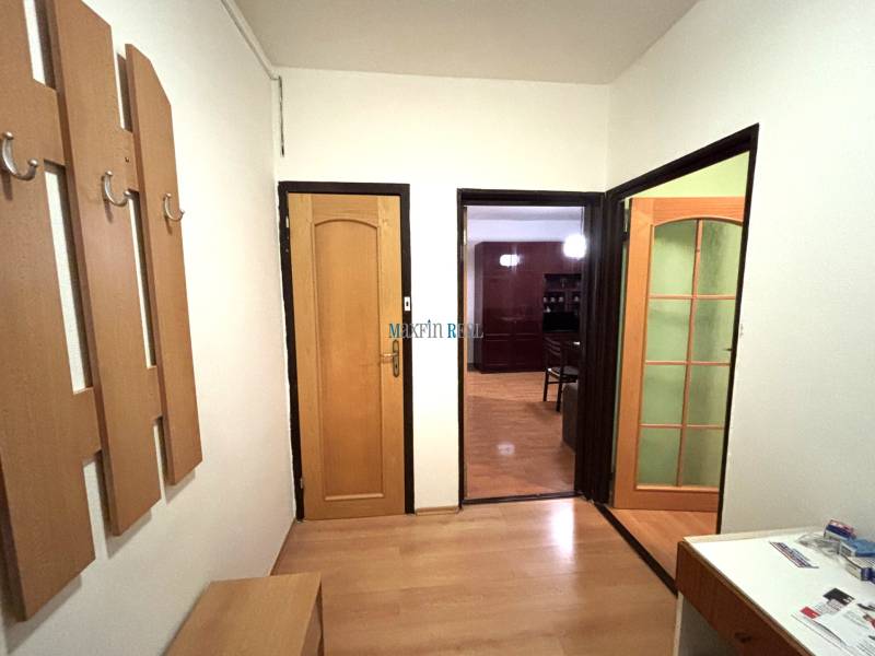 A two-room apartment with a wood-patterned floor, a coat rack wall, and open doors to a room.