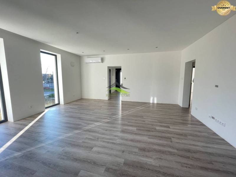 A spacious living room in a family house with wood-patterned flooring, large windows, and air conditioning.