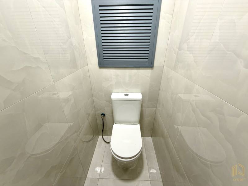 A toilet in a studio apartment with white tiles and a gray ventilation window.