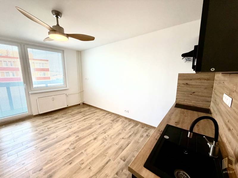 Studio apartment with a fan, kitchenette, and wood-patterned flooring.
