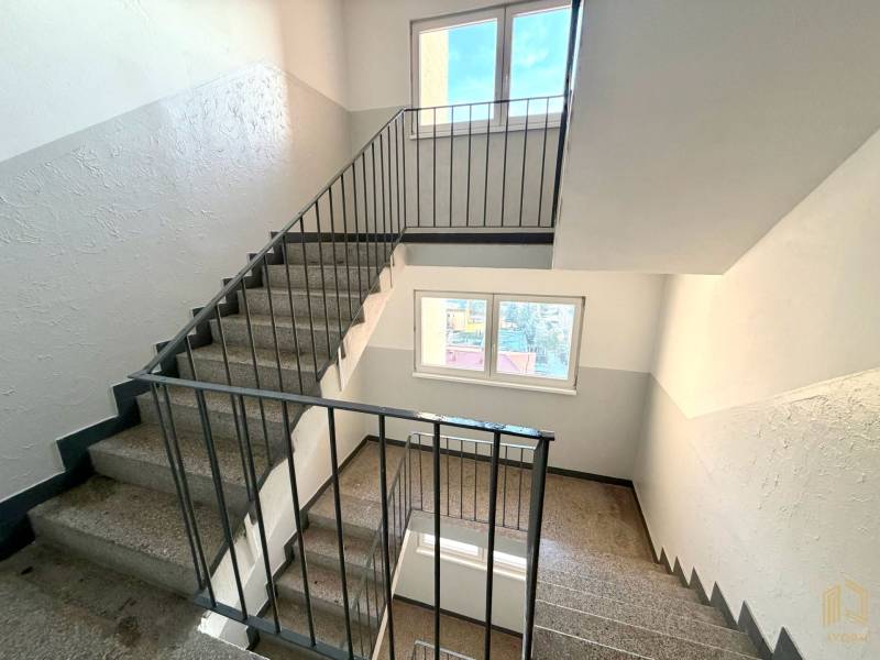 A staircase in a studio apartment with windows, white walls, and a metal railing.