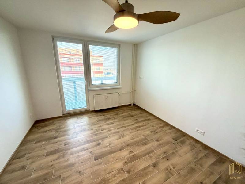 A room in a studio apartment with a wood-patterned floor, a ceiling fan, and a window with a view.