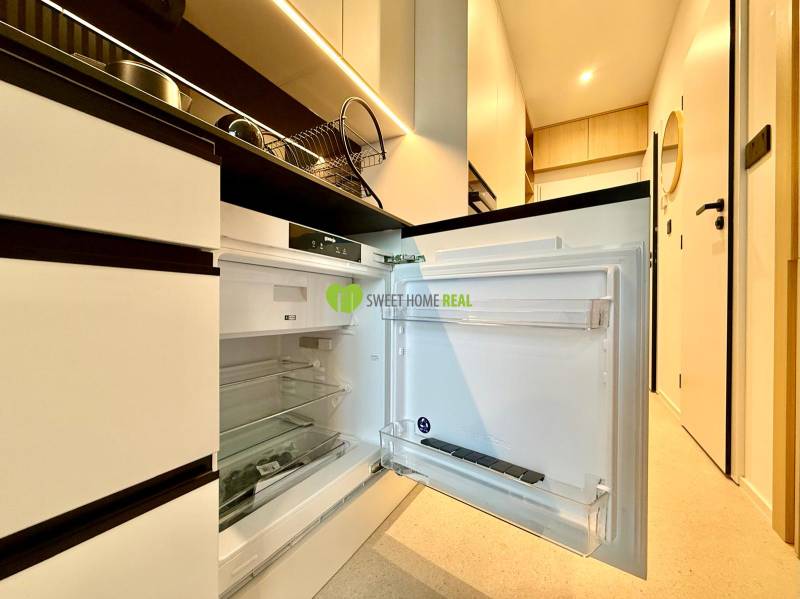 Kitchen area of a studio apartment with an open refrigerator and light-colored furniture.