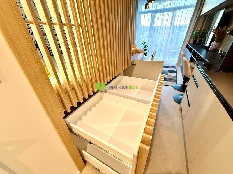 Interior of a 1-room apartment with wood decor, drawers, and a large window with white curtains.