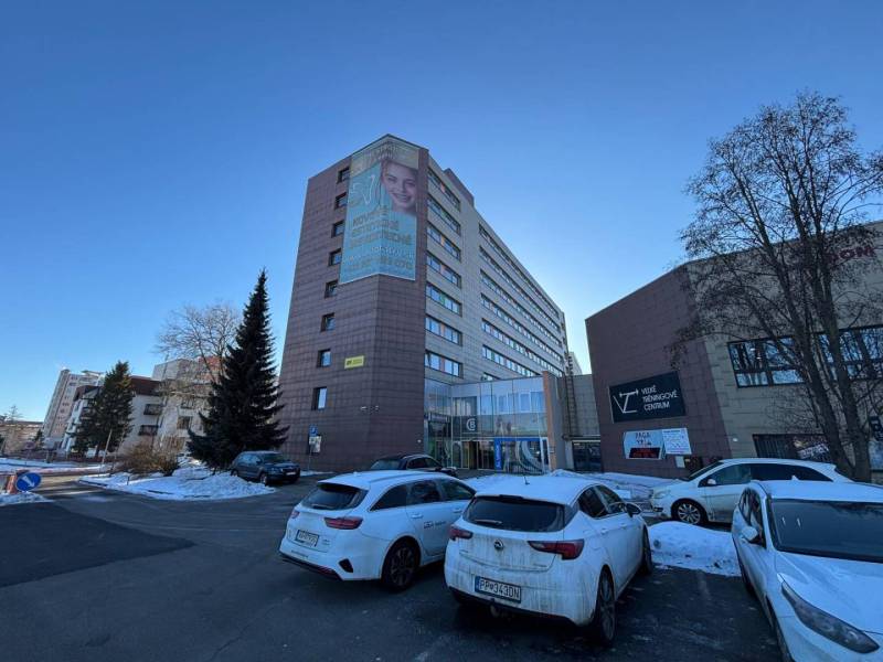 Office building in Poprad, snow-covered cars and road with blue sky.